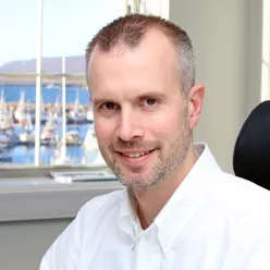 En smilende mann antrukket i hvitt med hav og båter i bakgrunnen